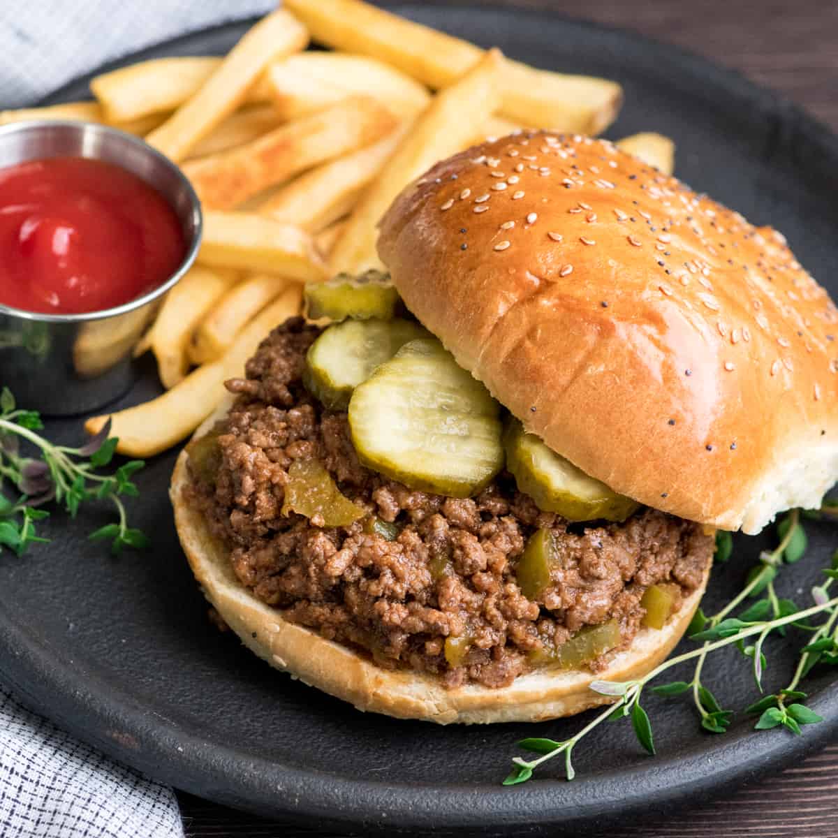 healthy sloppy joes recipe on a bun with pickles on a round black plate with ketchup and french fries and fresh thyme