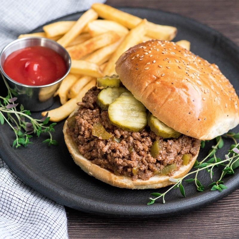  Draufsicht auf gesundes Sloppy Joes Rezept auf einem Brötchen mit Gurken auf einem runden schwarzen Teller mit Ketchup und Pommes Frites und frischem Thymian