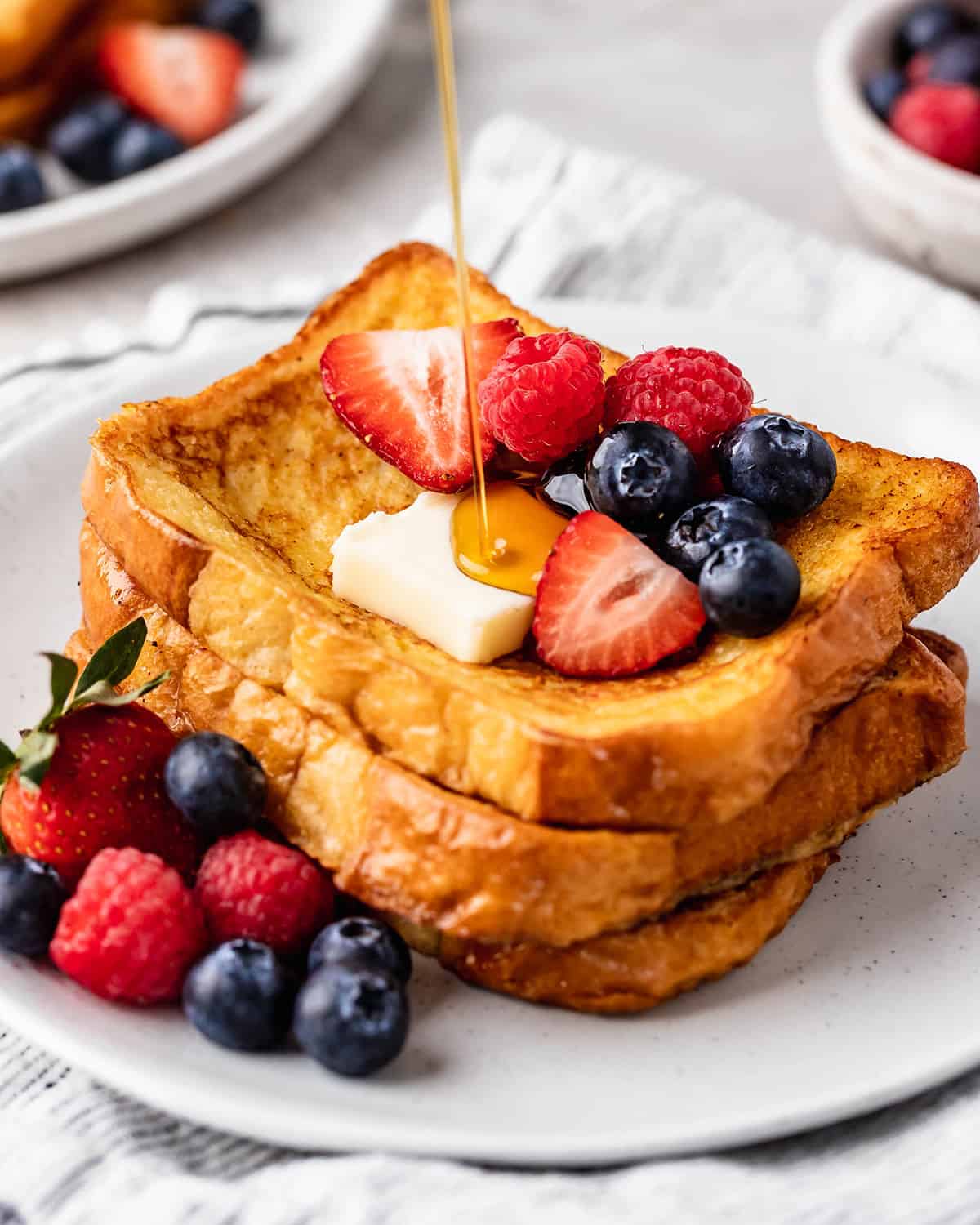 syrup being poured onto a stack of 3 pieces of French Toast with butter and berries