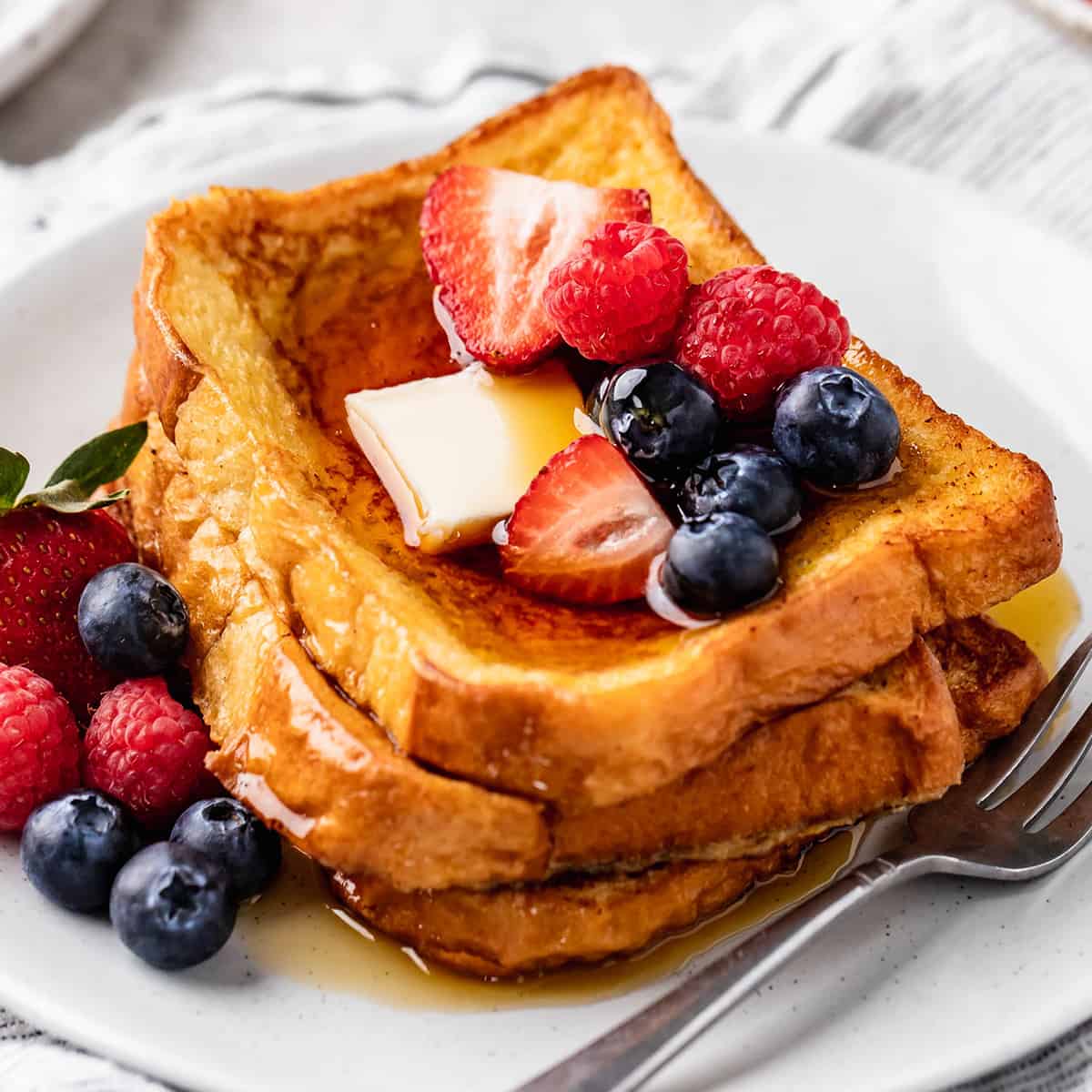 stack of three pieces of french toast on a plate with butter, syrup and berries
