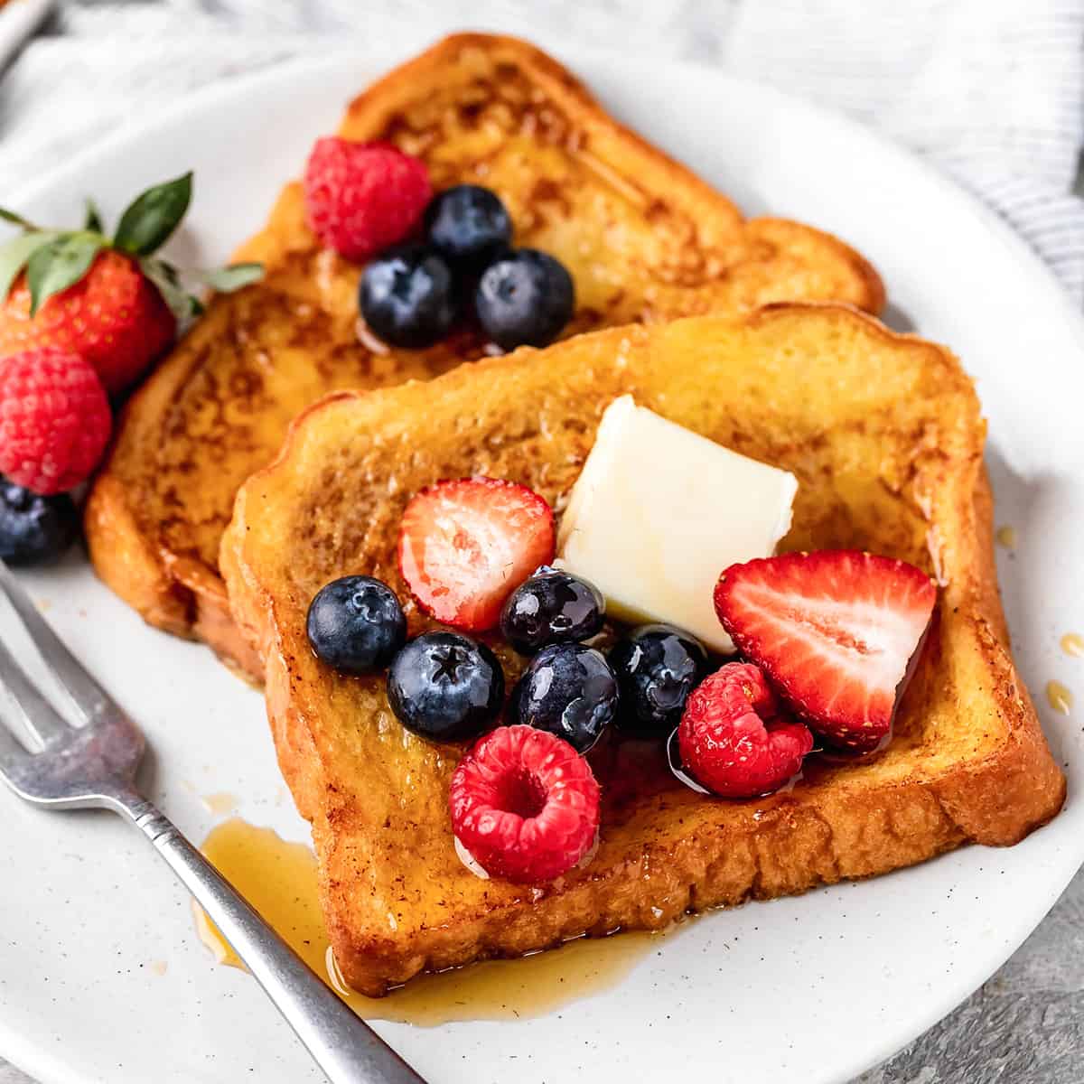 two pieces of French Toast on a plate with butter, syrup and berries 