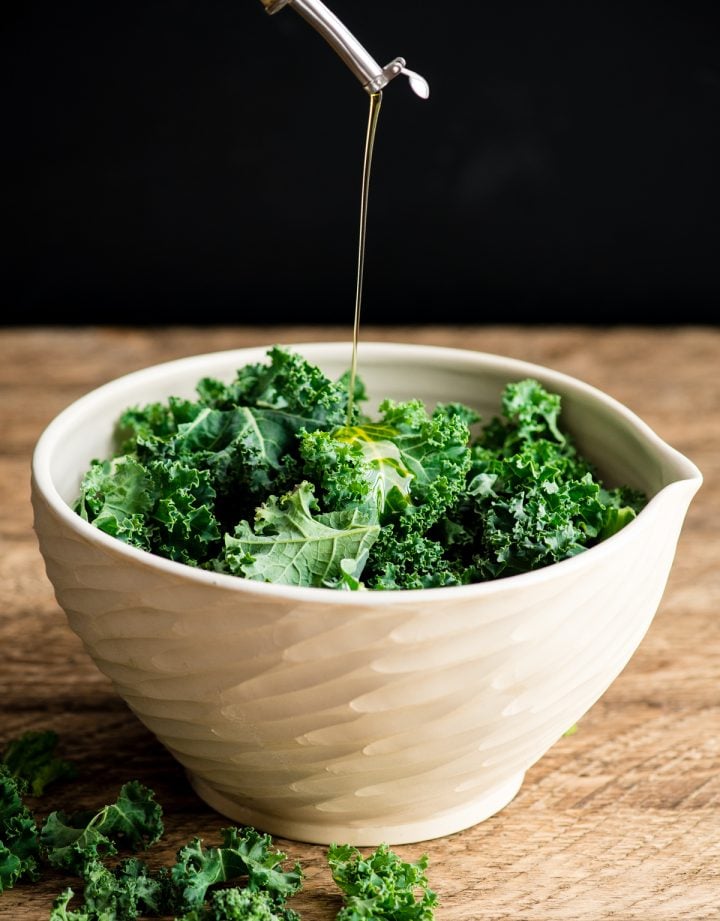 Front view of olive oil being poured into a bowl of chopped kale leaves making baked kale chips