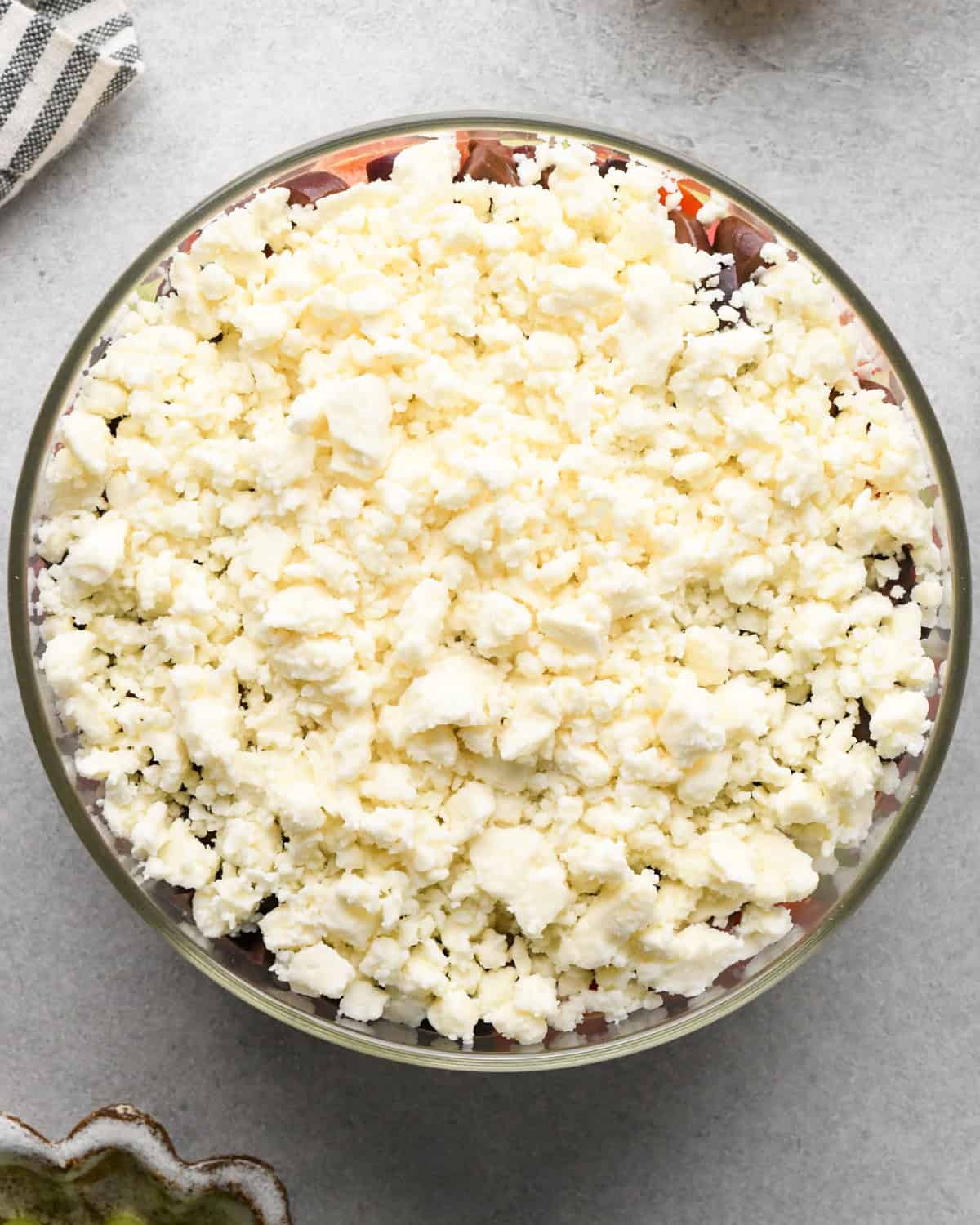 assembling layered greek dip, feta cheese spread on top of the kalamata olive layer in a glass bowl, on a gray surface.