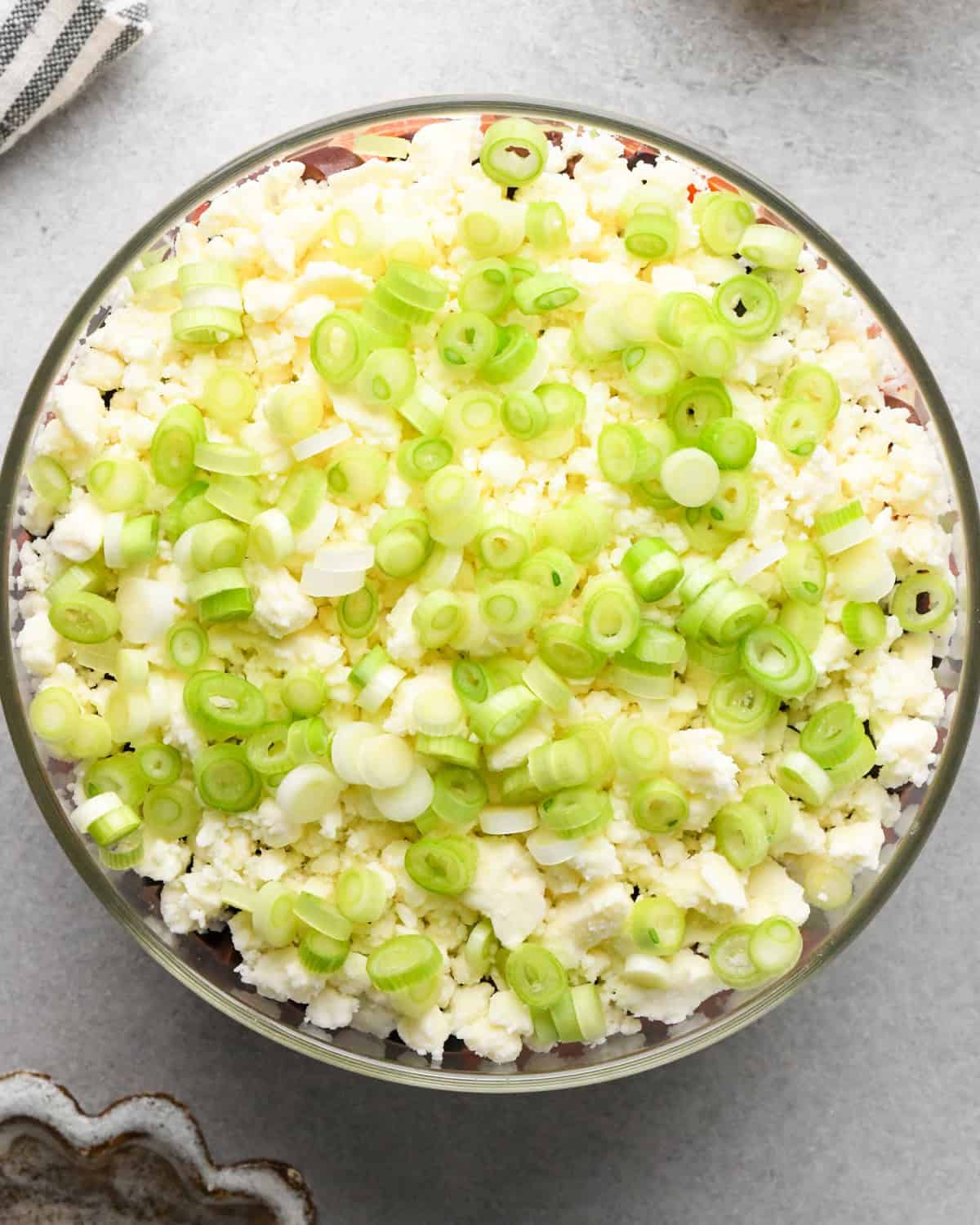 assembling layered greek dip, green onions spread on top of the feta cheese layer in a glass bowl, on a gray surface.