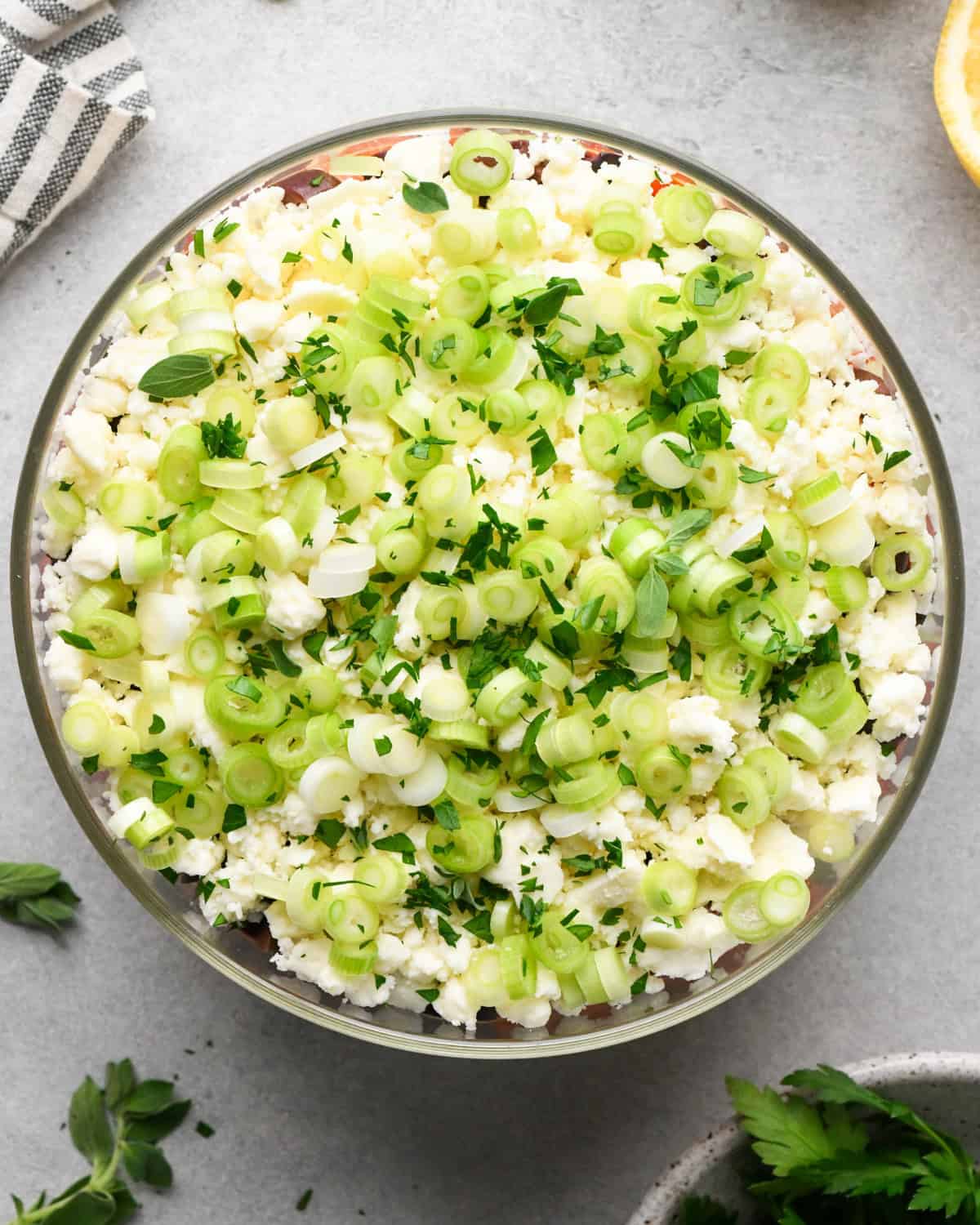 assembling layered greek dip, fresh herbs sprinkled on top of the green onions in a glass bowl, on a gray surface.