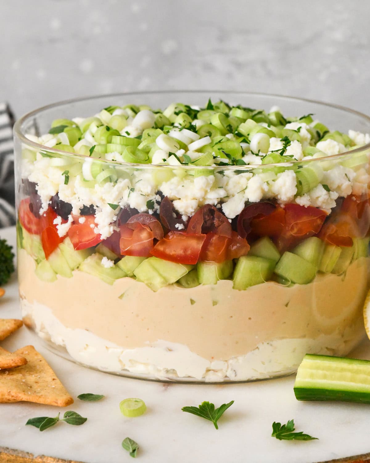 layered Greek dip in a clear glass dish, showing the layers of cream cheese, hummus, cucumbers, tomatoes, olives, feta cheese, and green onions, surrounded by fresh vegetables and crackers for dipping.