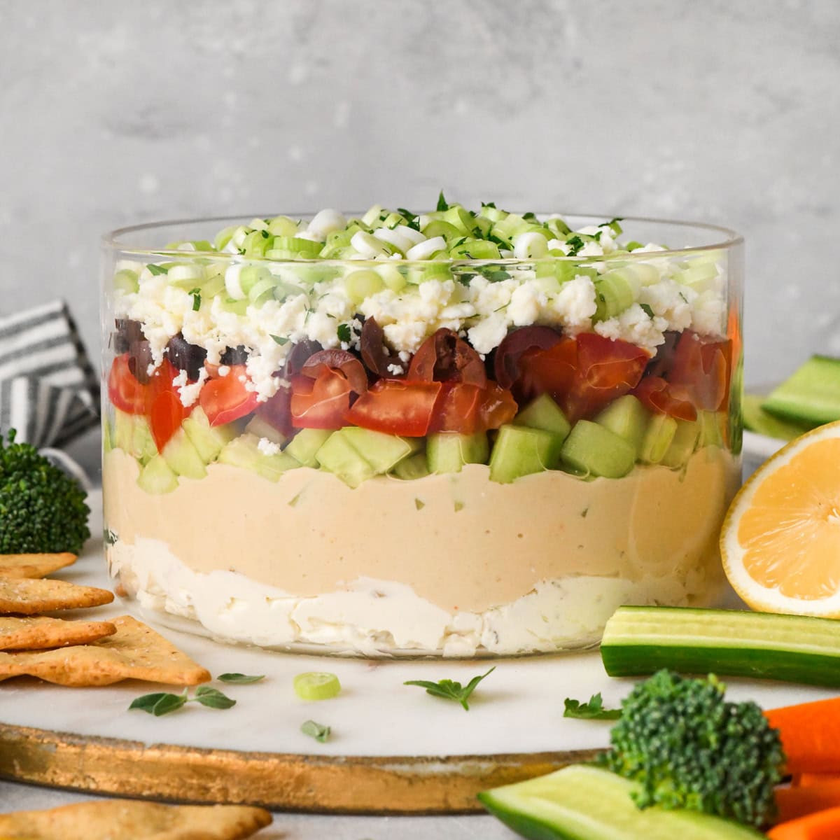 This Layered Greek Dip Recipe is the perfect healthier appetizer to serve when you're entertaining this summer! It's always the hit of the party (or BBQ)! It's gluten-free and only takes 10 minutes of prep time! #recipe #appetizer #healthy #glutenfree #bbq #dip #Greekdip #layereddip layered Greek dip in a clear glass dish, showing the layers of cream cheese, hummus, cucumbers, tomatoes, olives, feta cheese, and green onions, surrounded by fresh vegetables and crackers for dipping.