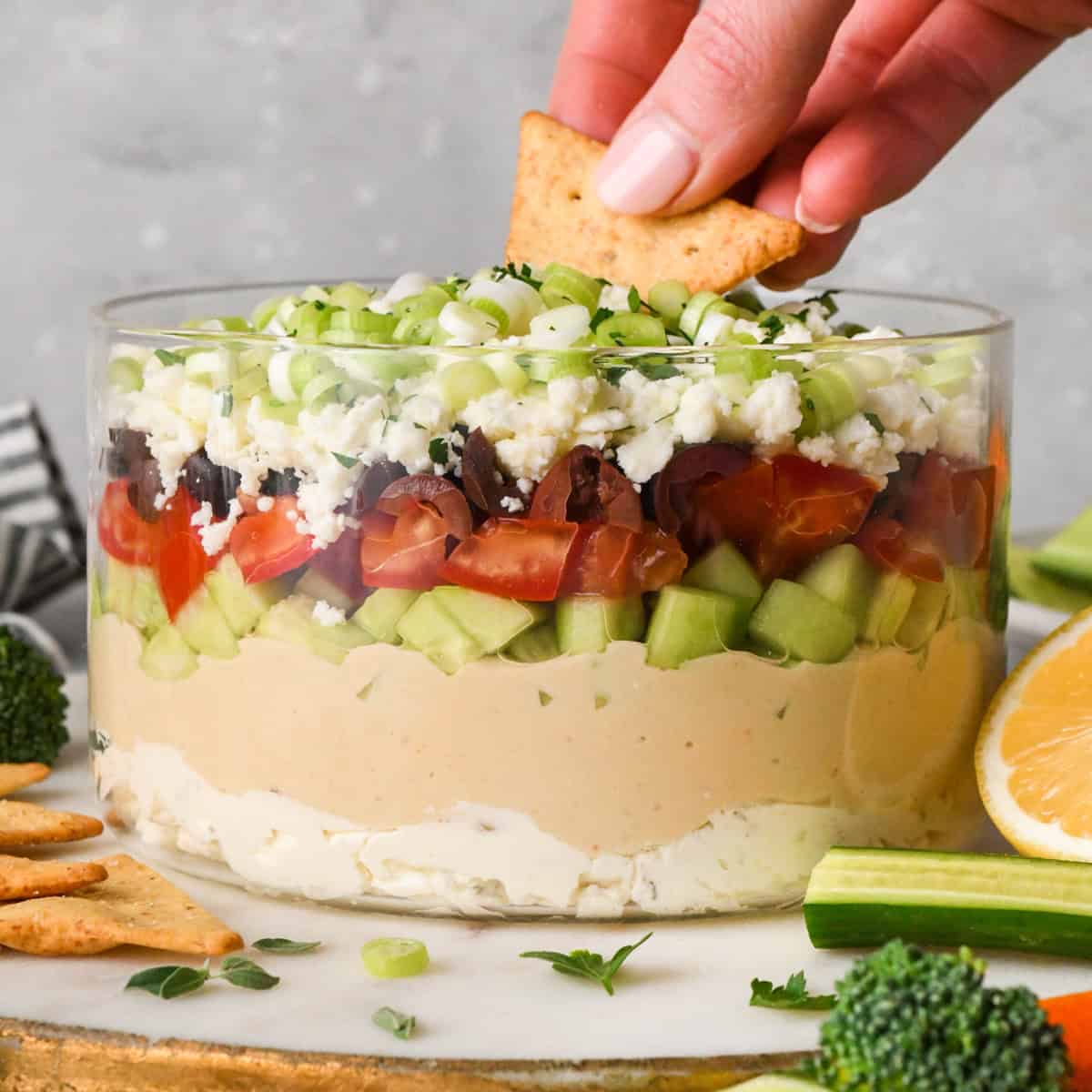 a hand dipping a pita chip into layered Greek dip in a clear glass dish, showing the layers of cream cheese, hummus, cucumbers, tomatoes, olives, feta cheese, and green onions, surrounded by fresh vegetables and crackers for dipping.