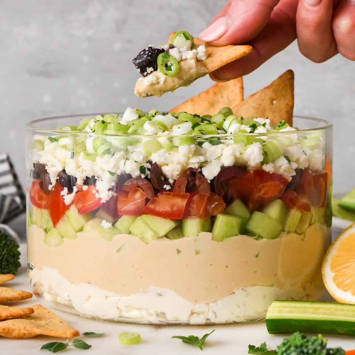 a hand holding a pita chip that has been dipped in greek dip over layered Greek dip in a clear glass dish, showing the layers of cream cheese, hummus, cucumbers, tomatoes, olives, feta cheese, and green onions, surrounded by fresh vegetables and crackers for dipping.