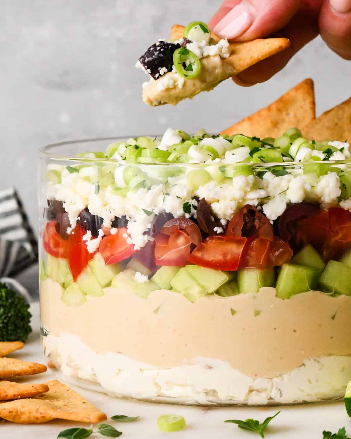 a hand holding a pita chip that has been dipped in greek dip over layered Greek dip in a clear glass dish, showing the layers of cream cheese, hummus, cucumbers, tomatoes, olives, feta cheese, and green onions, surrounded by fresh vegetables and crackers for dipping.