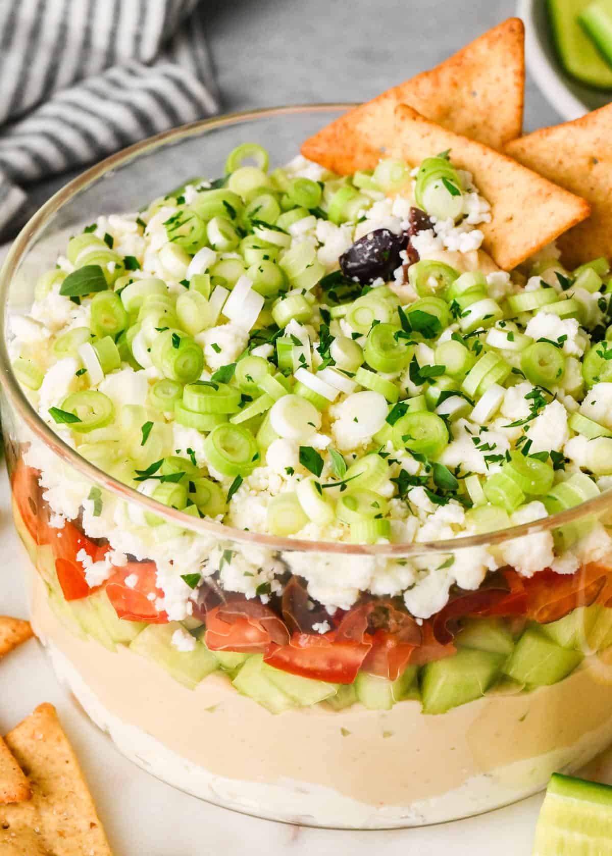 3 pita chips in layered greek dip  in a clear glass dish, showing the layers of cream cheese, hummus, cucumbers, tomatoes, olives, feta cheese, and green onions, surrounded by fresh vegetables and crackers for dipping.