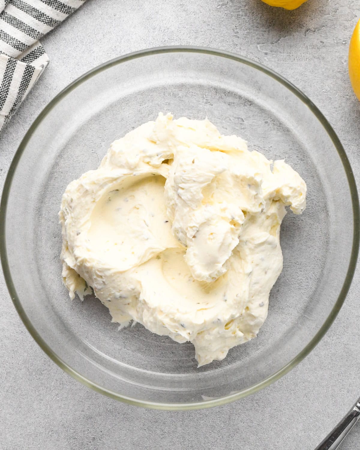 herbed cream cheese for the bottom layer of this greek dip recipe in the bottom of a glass bowl before spreading, on a gray surface.