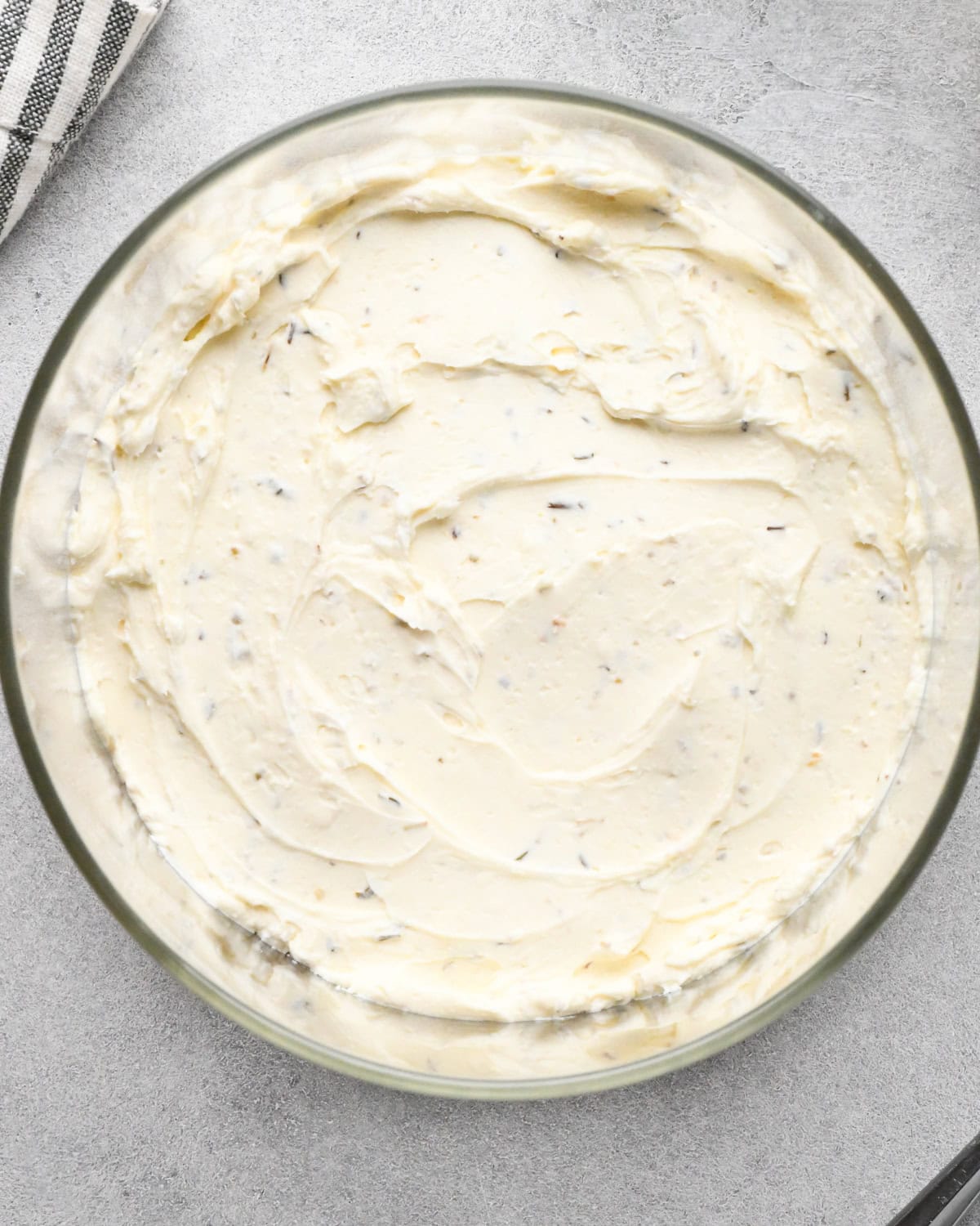assembling layered greek dip, the herbed cream cheese layer evenly spread in the bottom of a glass bowl, on a gray surface.
