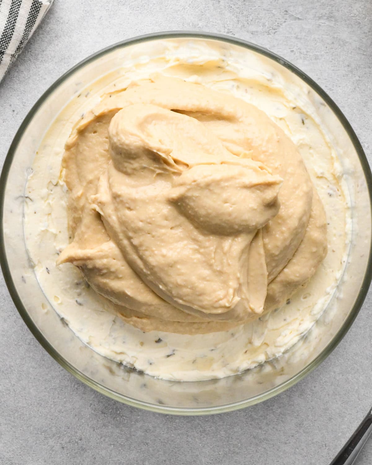 assembling layered greek dip, hummus on top of the herbed cream cheese layer before spreading in a glass bowl, on a gray surface.