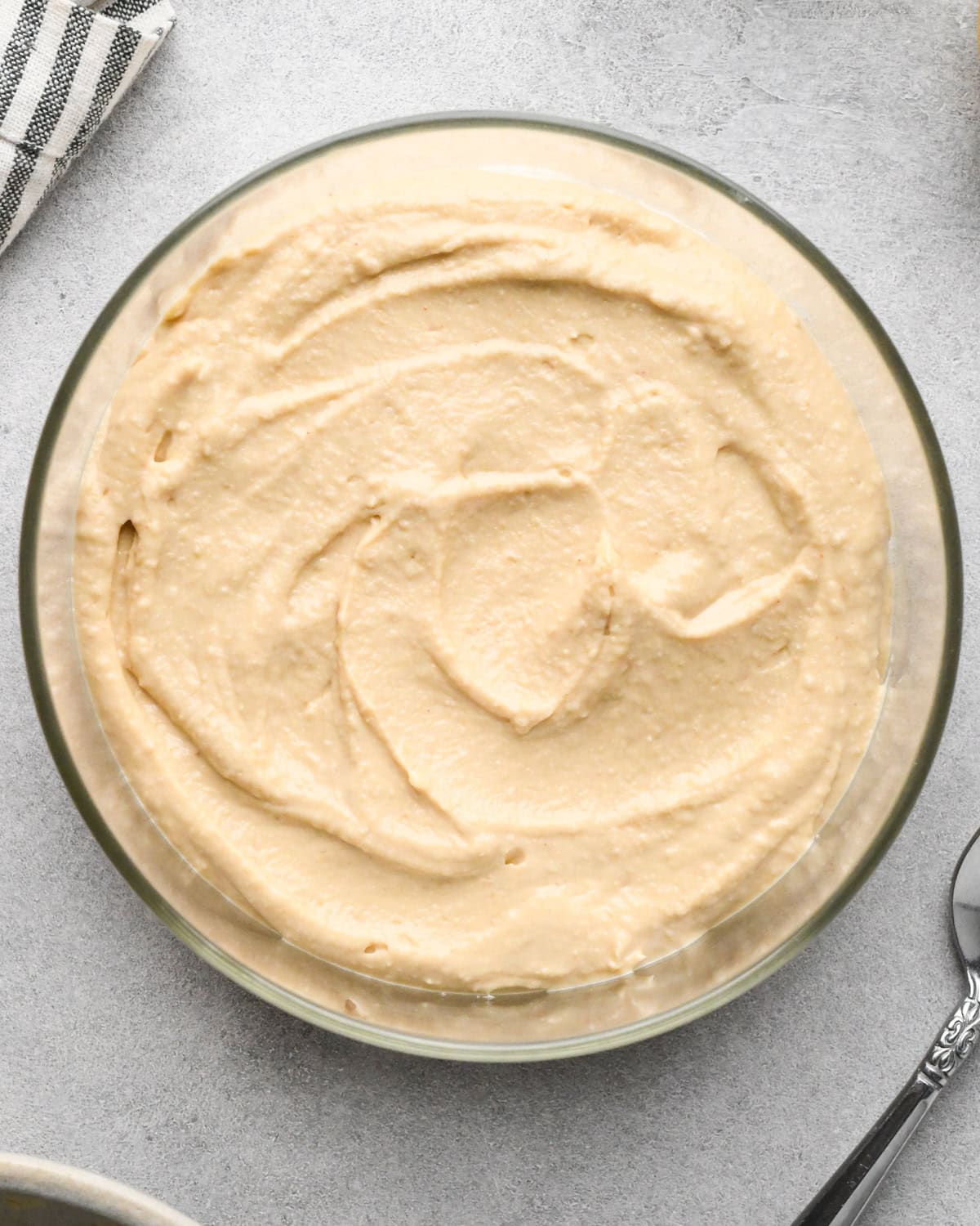 assembling layered greek dip, the hummus layer evenly spread on top of the cream cheese layer in a glass bowl, on a gray surface.