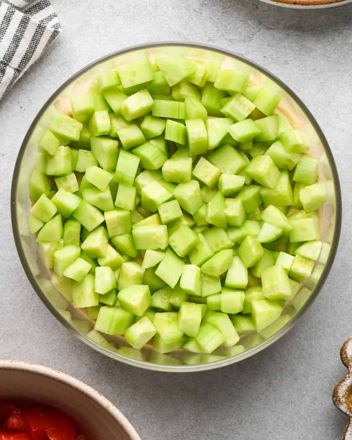 assembling layered greek dip, cucumbers spread on top of the hummus layer in a glass bowl, on a gray surface.