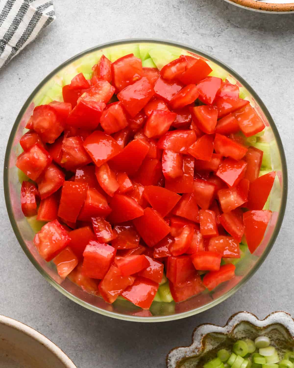 assembling layered greek dip, tomatoes spread on top of the cucumber layer in a glass bowl, on a gray surface.