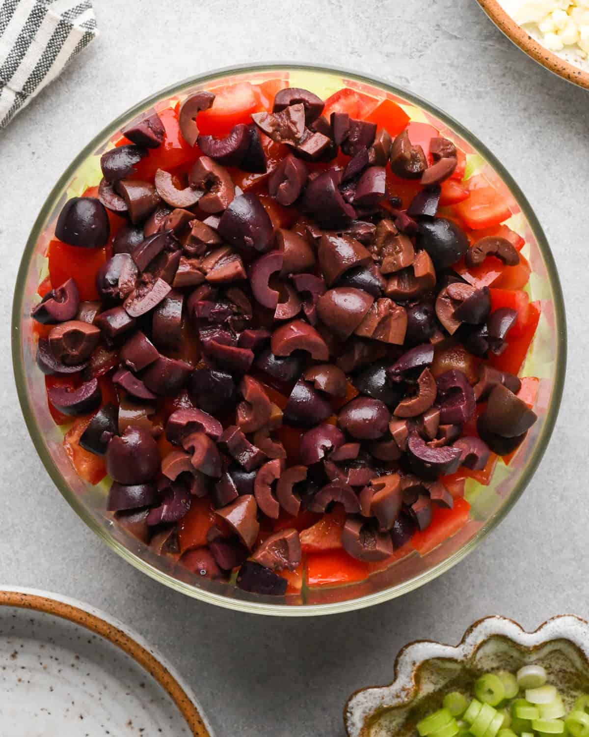 assembling layered greek dip, kalamata olives spread on top of the tomato layer in a glass bowl, on a gray surface.