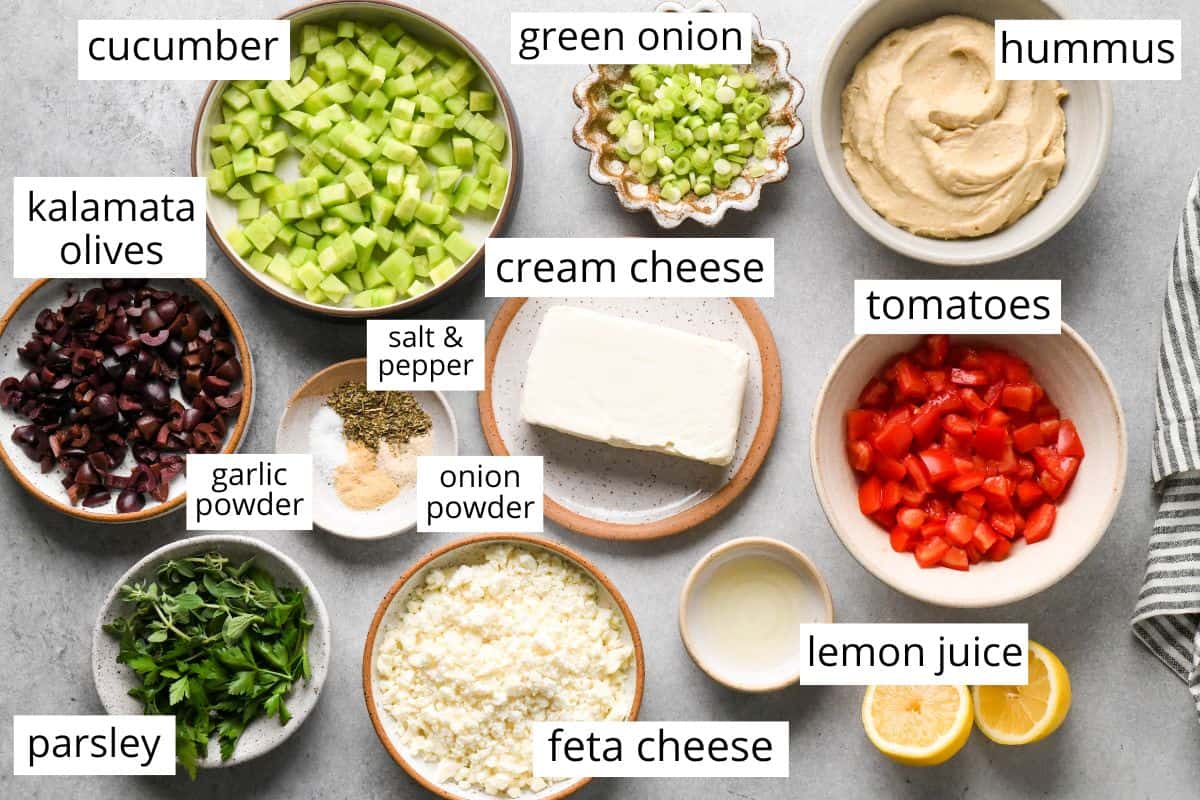 overhead photo of the labeled ingredients in this layered greek dip recipe - like cream cheese, cucumbers, olives, hummus, tomatoes, feta cheese and more. 