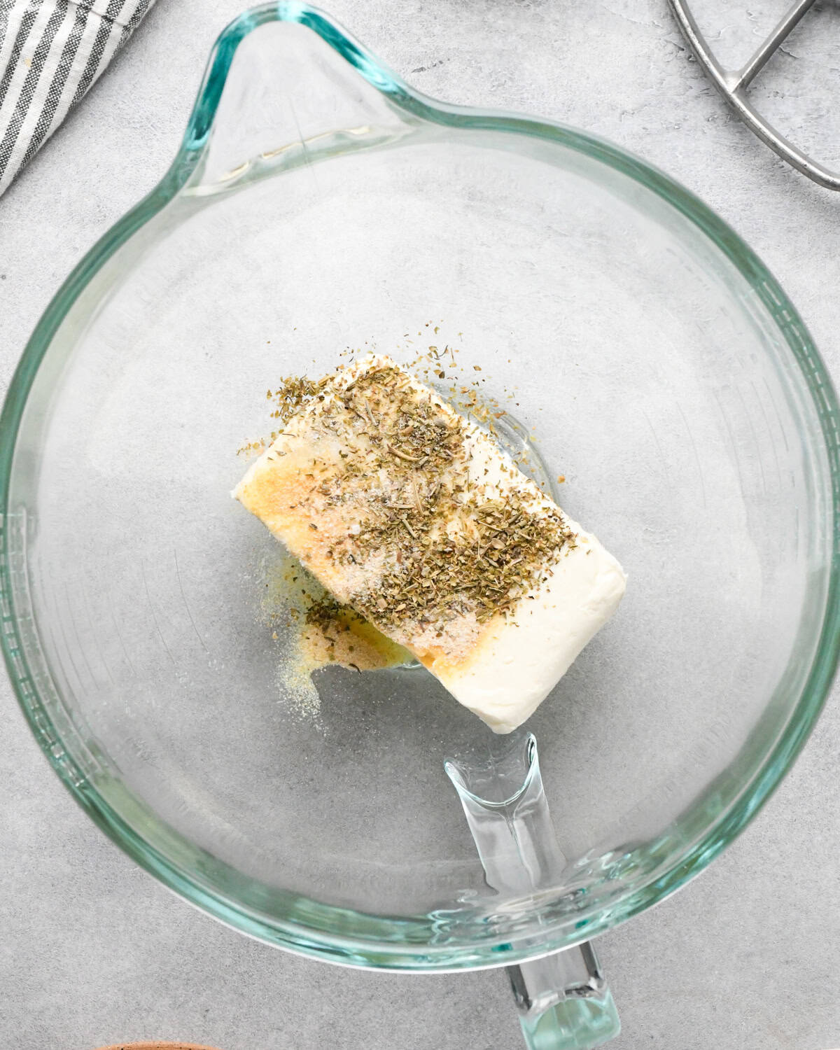 a block of cream cheese topped with dried herbs and seasonings in a glass mixing bowl, ready to be made into the base for this layered Greek dip recipe