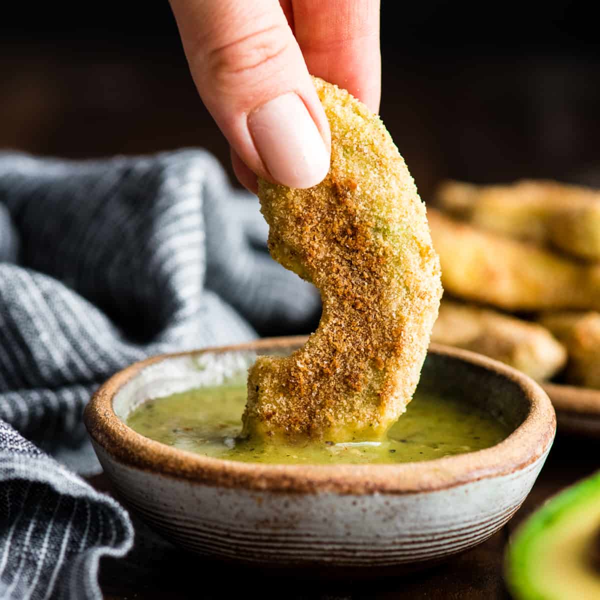 front view of a hand dipping an avocado fry into sauce 