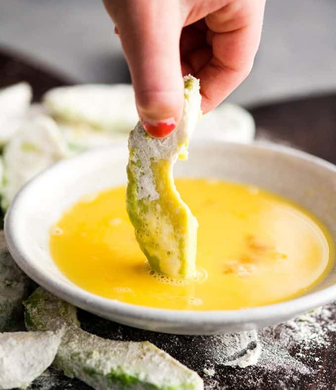 Vorderansicht einer Hand, die bei der Herstellung von gebackenen Avocado-Pommes eine mit Mehl überzogene Avocado-Scheibe in eine geschlagene Ei-Mischung taucht