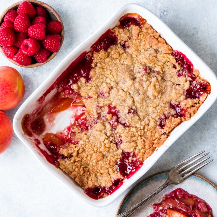 Overhead view of a Raspberry Peach Crisp in a baking dish with a serving scooped out on a plate nearby