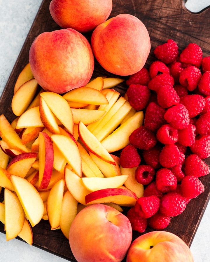 This easy Raspberry Peach Crisp is one of our favorite dessert recipes! The buttery crumb topping paired with the sweet peaches and tart raspberries is absolute summer perfection! It's easily made gluten-free! #peach #raspberry #crisp #peachcrisp #raspberrycrisp #dessert #summer #berrycrisp #glutenfree Overhead view of slices peaches and full raspberries on a cutting board in the making of Raspberry Peach Crisp