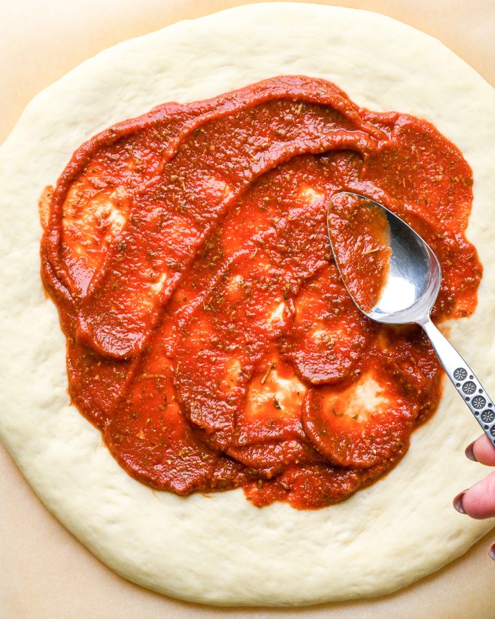 pizza sauce being spread onto pizza dough with a spoon