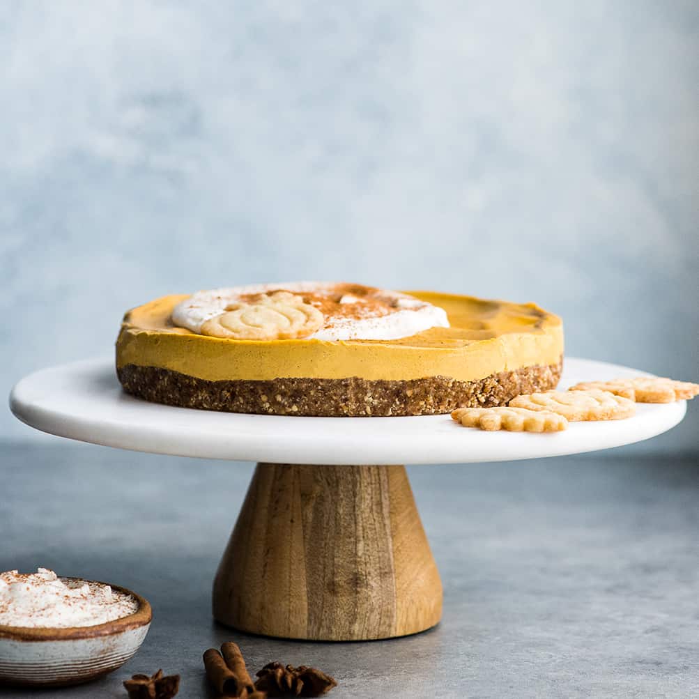 Front view of a full No-Bake Vegan Pumpkin Cheesecake on a cake stand topped with whipped cream and a sprinkle of cinnamon