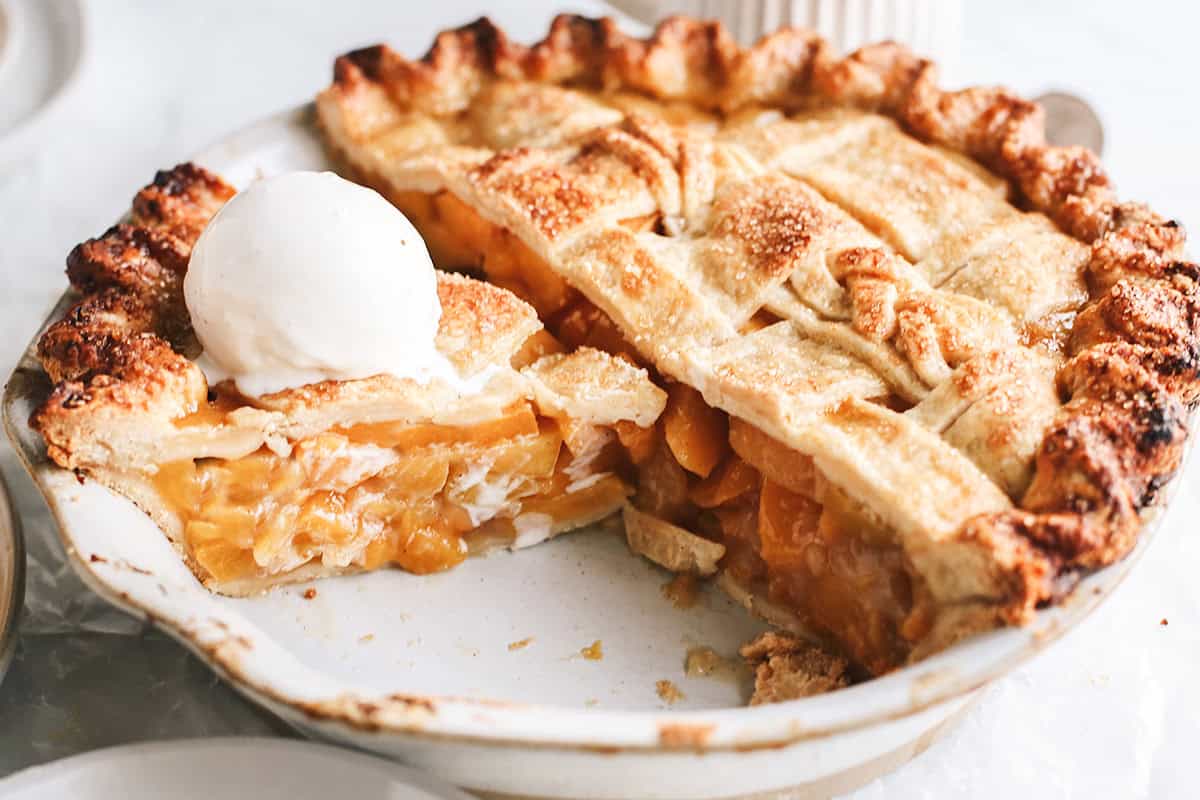 front view of a peach pie with a double Butter Pie Crust with a slice cut out and ice cream on top