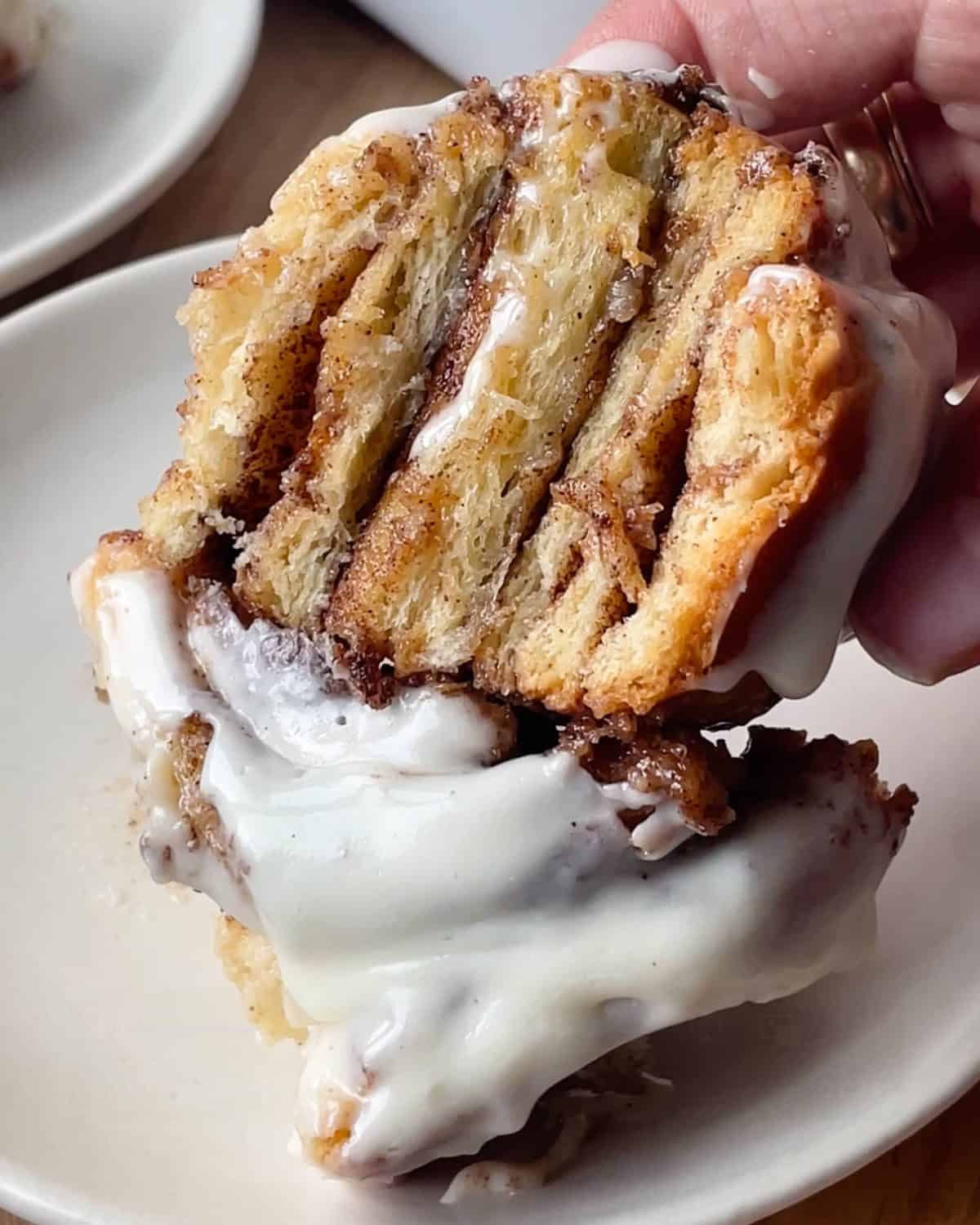 a hand holding half of a cinnamon roll over a plate with the other half of a cinnamon roll on it. You can see the gooey layers of cinnamon sugar inside the cinnamon roll and the cream cheese frosting on top.
