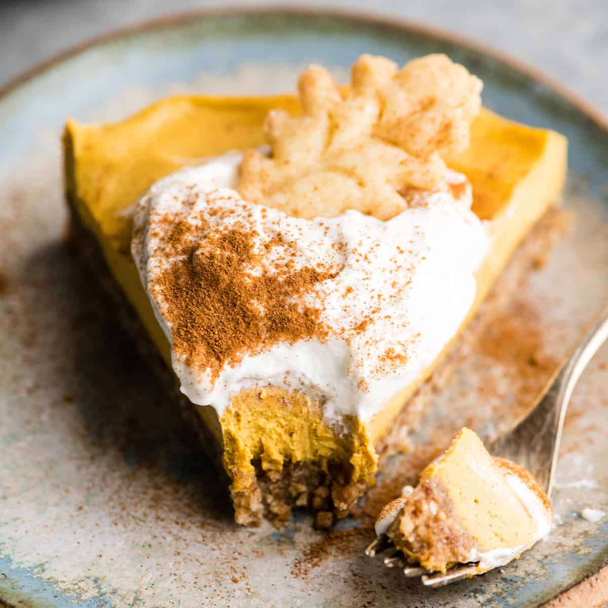 front view of a slice of No-Bake Vegan Pumpkin Cheesecake on a plate with a bite taken out of it on a fork sitting next to the slice of paleo Vegan Pumpkin Cheesecake