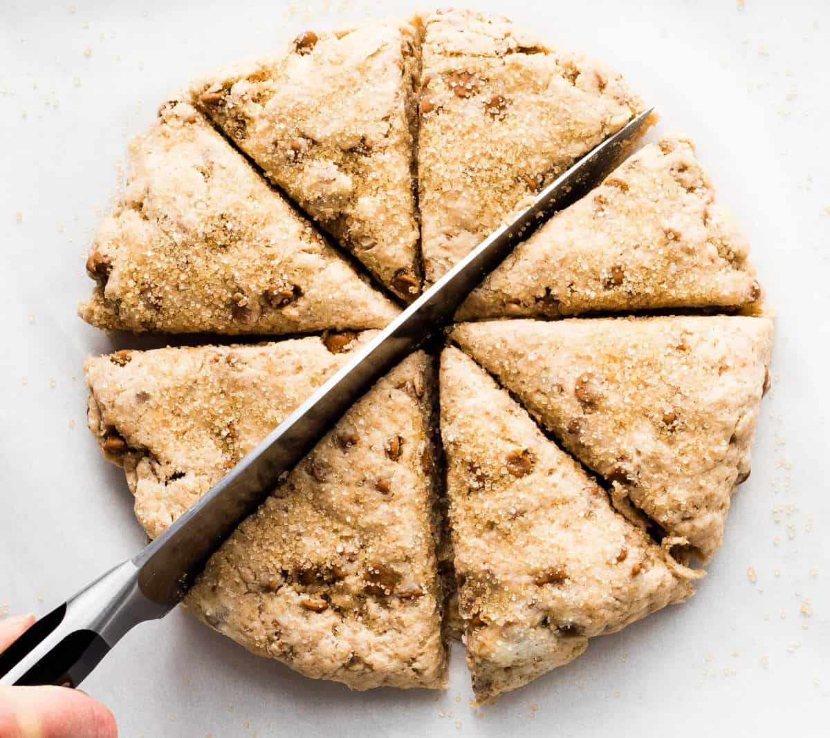 overhead view of a hand holding a knife cutting a circle of cinnamon chip scone dough into eight pieces. 