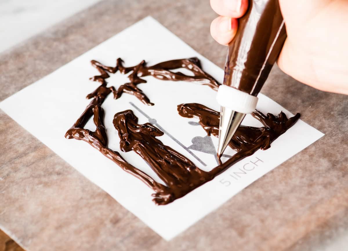 front view showing a hand using a decorating bag to trace the nativity cake topper with chocolate in the making of this Birthday Cake for Jesus