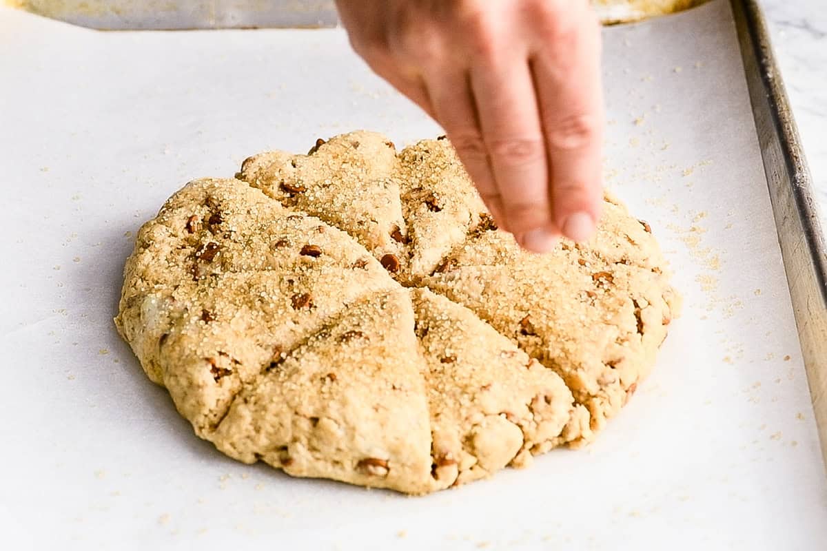 How to Make Cinnamon Scones - a hand sprinkling raw sugar over a disc of scone dough cut into 8 pieces