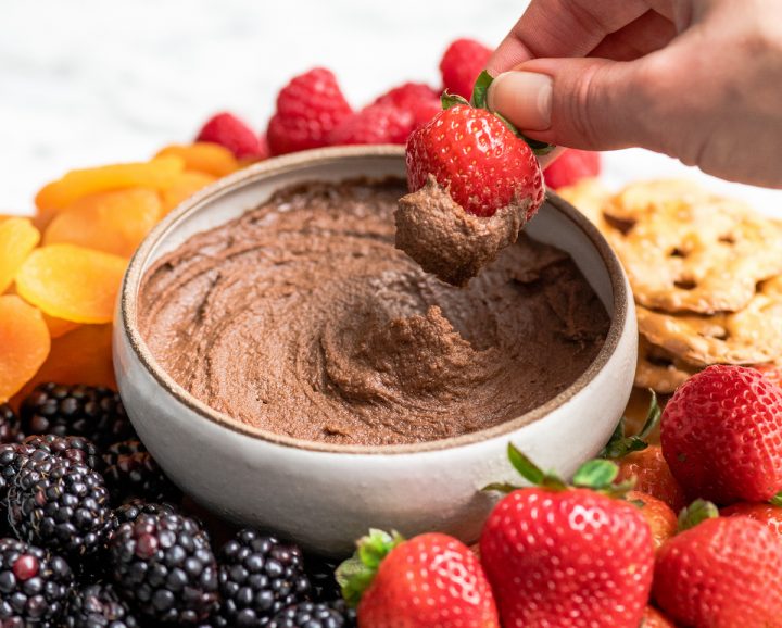 Front view of a hand dipping a strawberry into a bowl of Chocolate Hummus