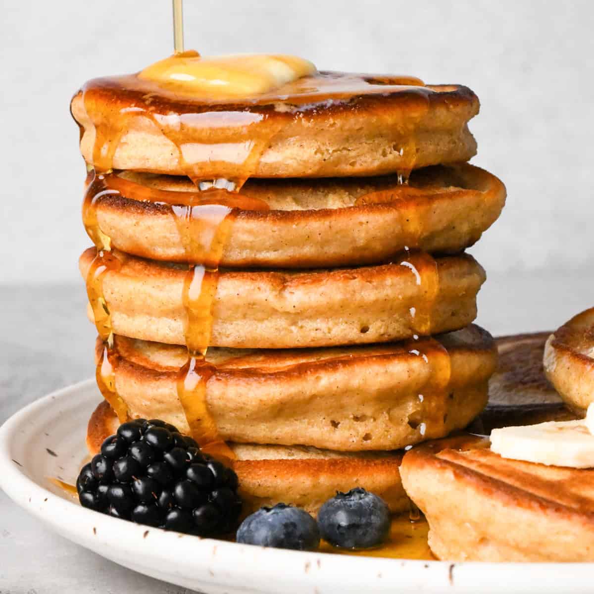 a stack of 5 greek yogurt pancakes on a white ceramic plate, there is melty butter on the top pancake and syrup is being poured over the stack. 