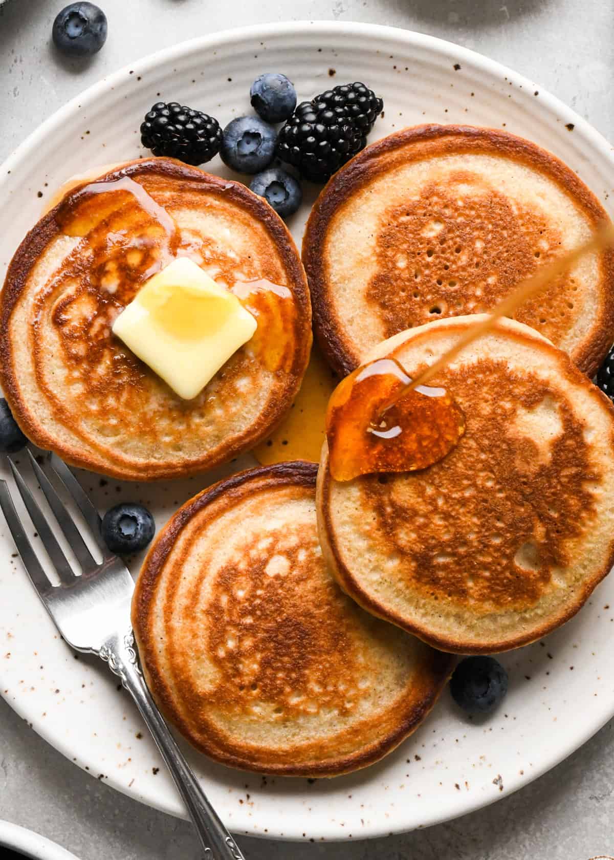 syrup being poured over 4 greek yogurt pancakes on a white ceramic plate, one has a square of butter on top and there are blueberries, blackberries and  fork on the plate. 