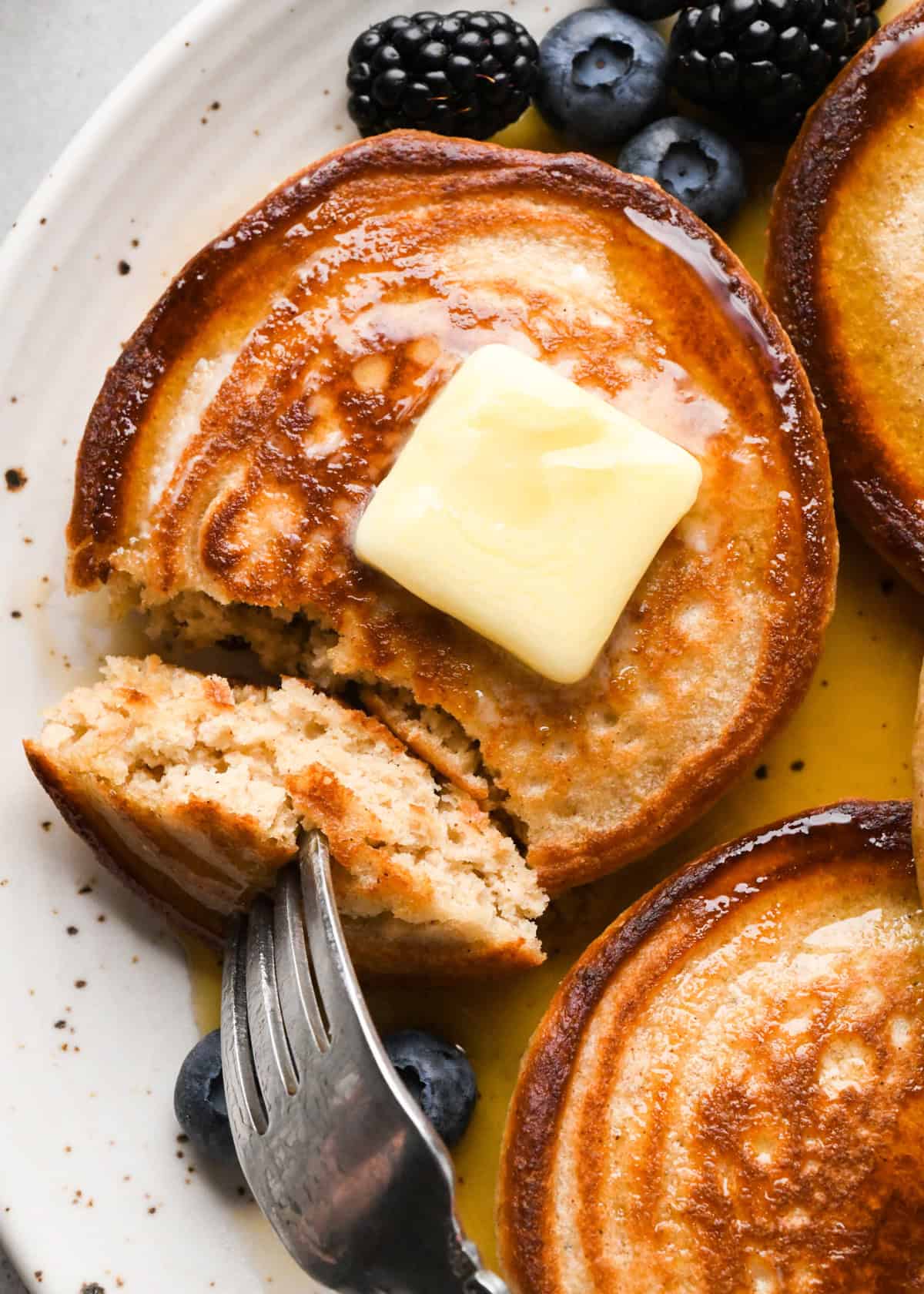 3 greek yogurt pancakes on a white ceramic plate with syrup and a square of melty butter is on the middle pancake, which also has a bite cut out of it on a fork showing the fluffy interior of the pancake. There are blueberries on the plate