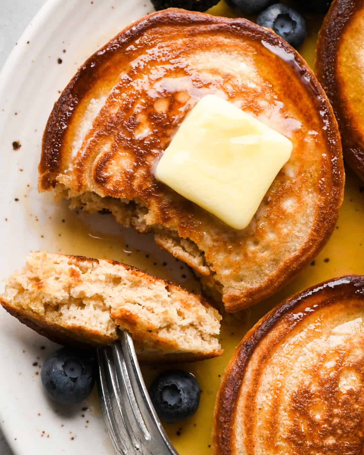 3 greek yogurt pancakes on a white ceramic plate with syrup and a square of melty butter is on the middle pancake, which also has a bite cut out of it on a fork showing the fluffy interior of the pancake. There are blueberries on the plate