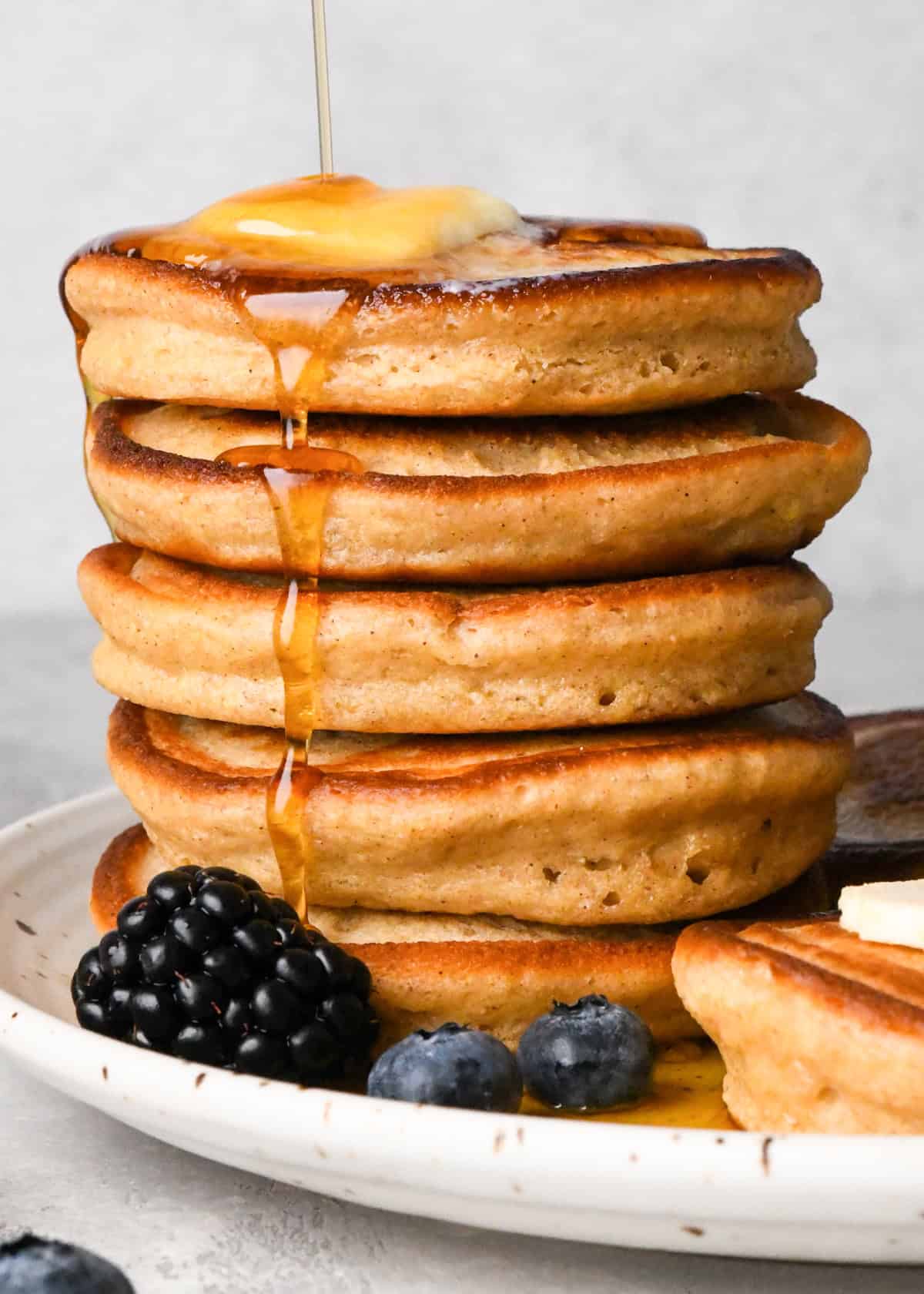 a stack of 5 greek yogurt pancakes on a white ceramic plate, there is melty butter on the top pancake and syrup is being poured over the stack. 