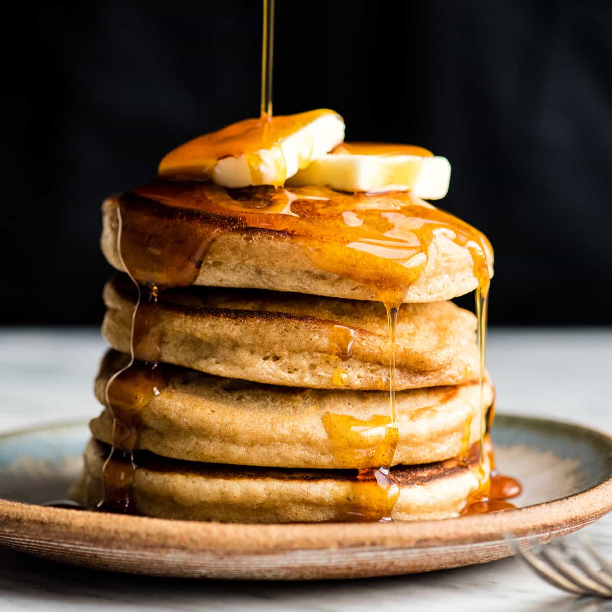 Front view of syrup being poured on a stack of four Greek Yogurt Pancakes with butter on top. 
