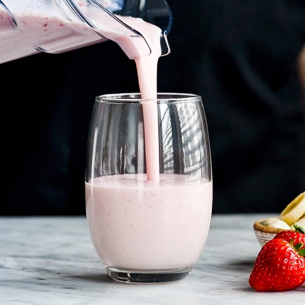 Front view of a strawberry banana smoothie being poured out of the blender into a glass cup. 