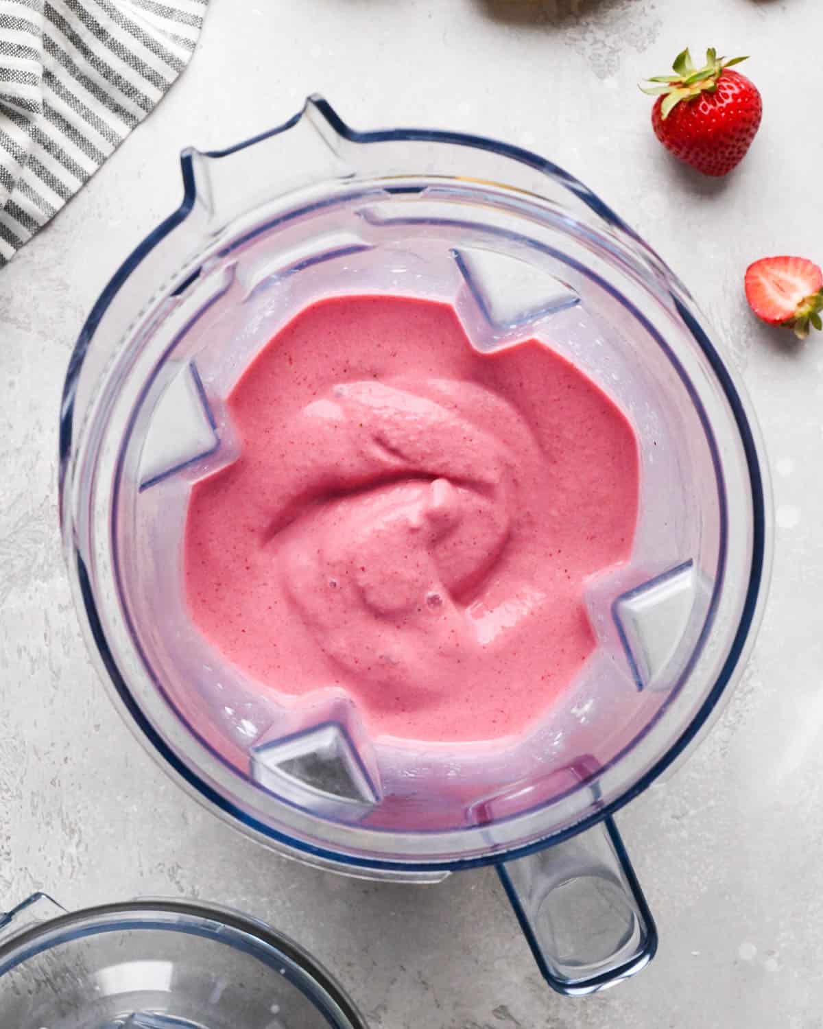 blended strawberry banana smoothie in a blending container surrounded by a striped towel and strawberries