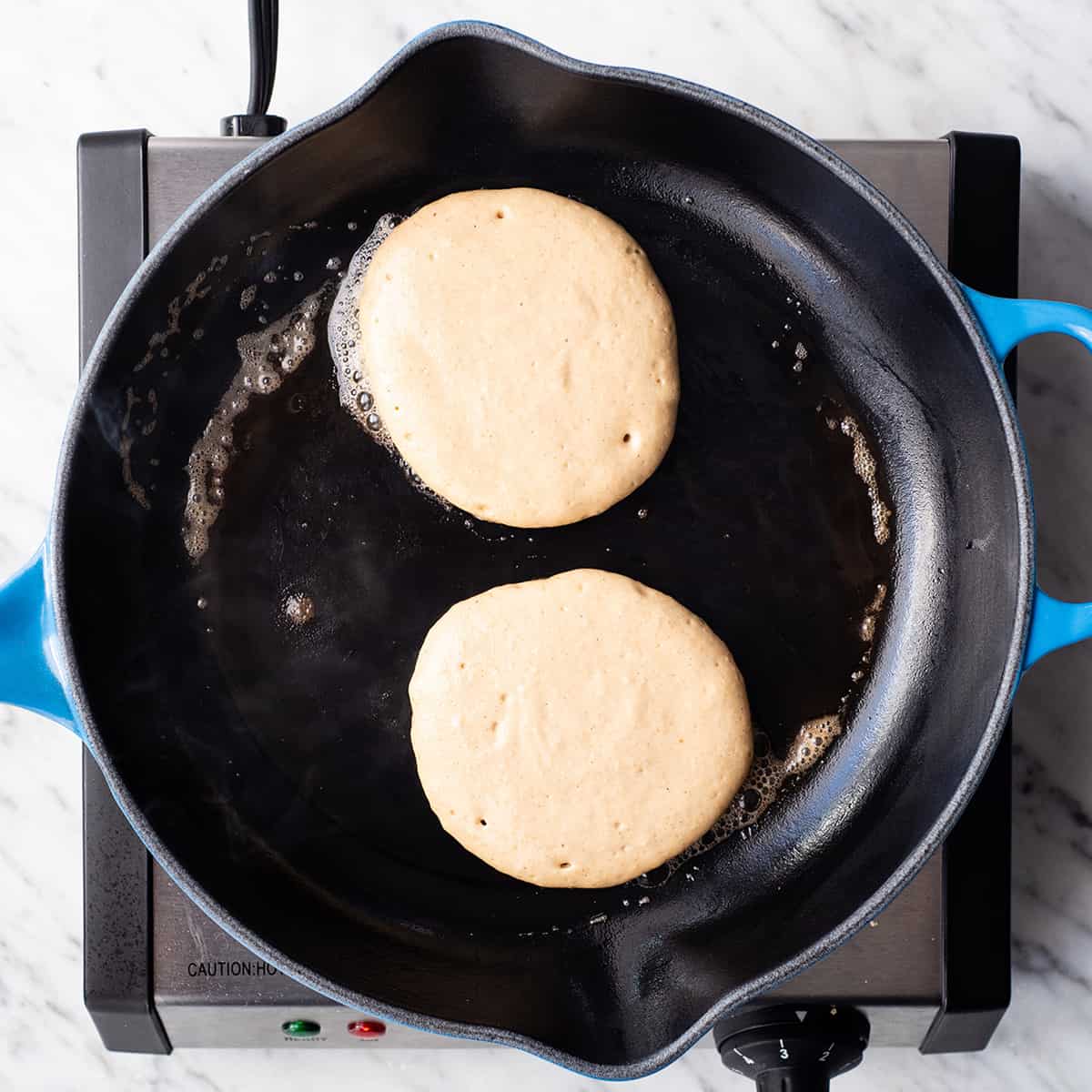 overhead view of two Greek Yogurt Pancakes cooking in a cast iron skillet before being flipped