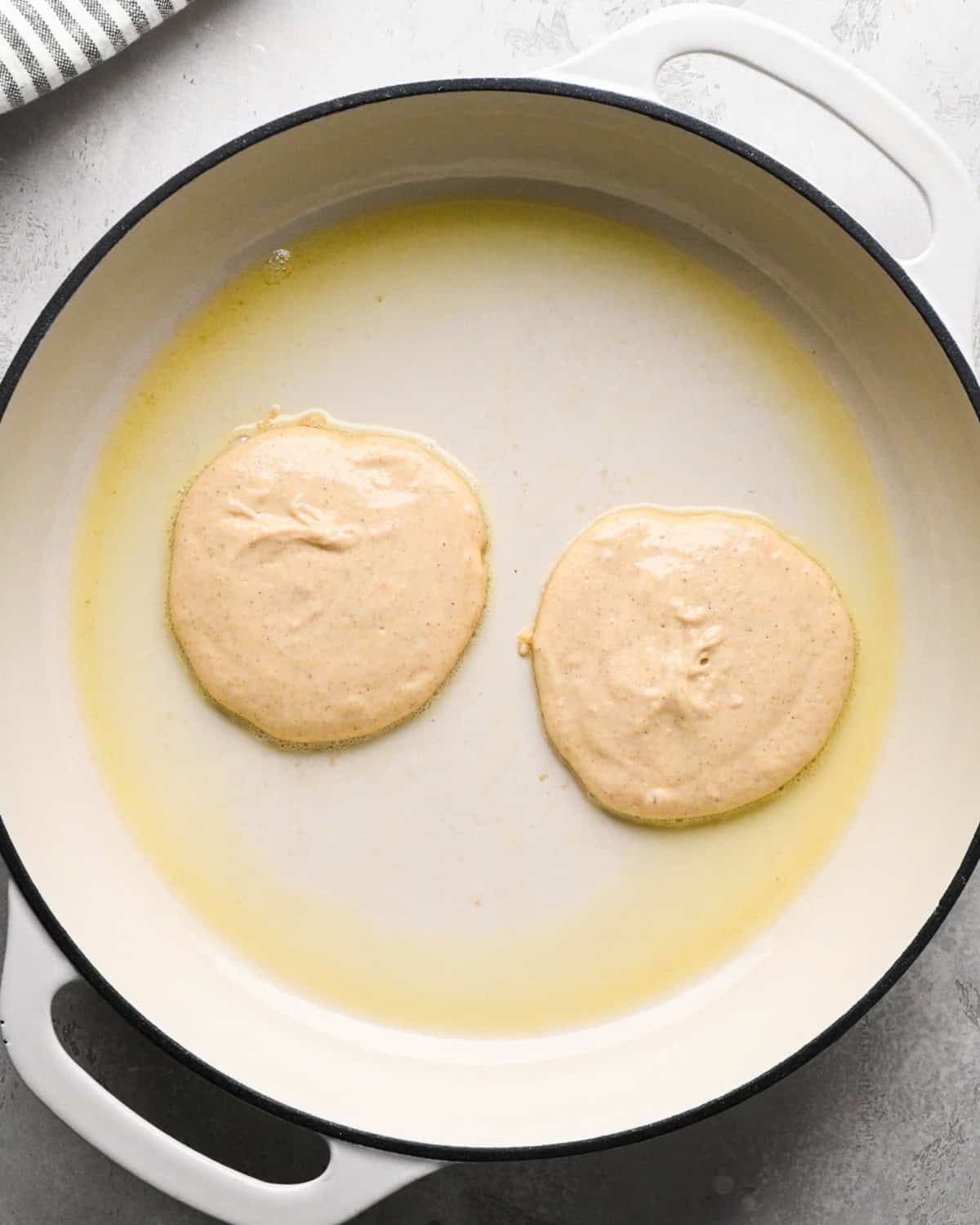 two greek yogurt pancakes at the beginning of cooking in a white buttered skillet