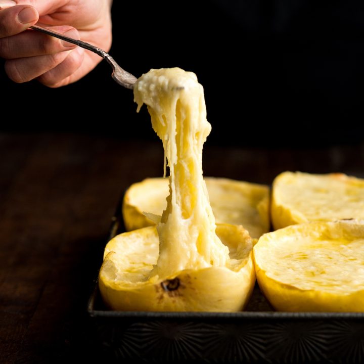 Front view of a hand holding a fork taking a bite of Spaghetti Squash Mac and Cheese out of one of four halves in a baking sheet