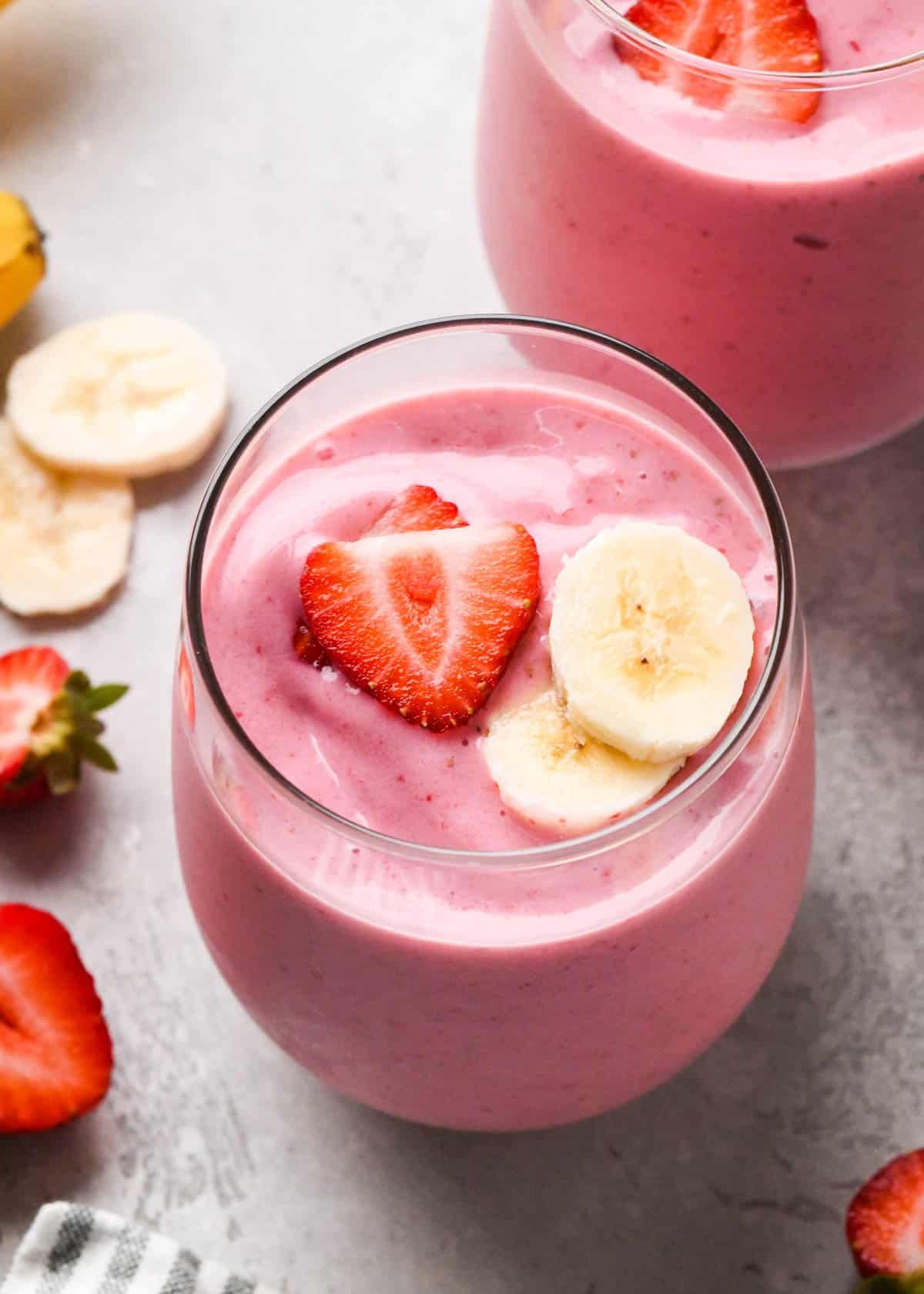 The BEST strawberry banana smoothie recipe ever! It's easy (ready in less than 5 minutes), healthy, and made with yogurt so it's full of protein! It's the perfect snack or breakfast! #smoothie #strawberrybananasmoothie #strawberry #banana #breakfast #snack #vitamix strawberry smoothie in a glass topped with two sliced bananas and two sliced strawberries, there is another smoothie in a glass, strawberries and bananas in the background