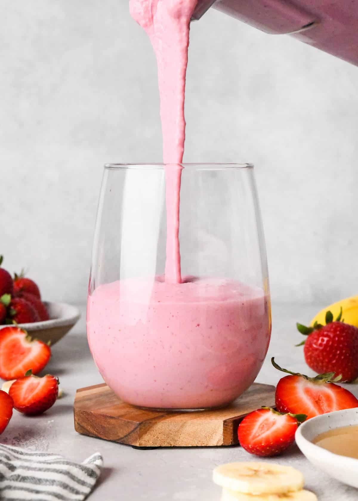 The BEST strawberry banana smoothie recipe ever! It's easy (ready in less than 5 minutes), healthy, and made with yogurt so it's full of protein! It's the perfect snack or breakfast! #smoothie #strawberrybananasmoothie #strawberry #banana #breakfast #snack #vitamix strawberry banana smoothie being poured from a blending container into a glass which is half full of the smoothie. Surrounded by strawberries and bananas the glass is on a wooden coaster